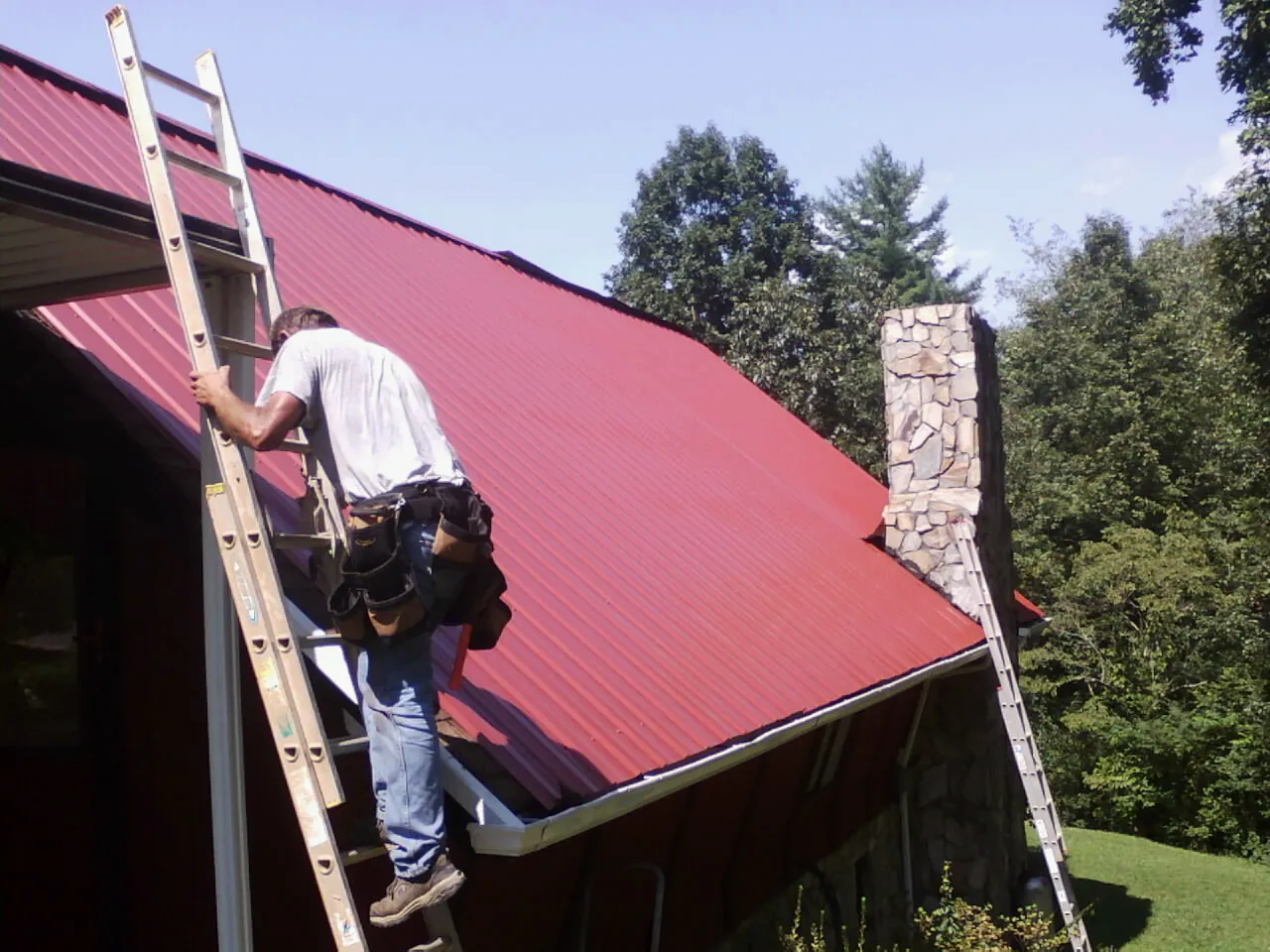 Roofer on a ladder beside a red metal roof and stone chimney — Soffit & Fascia Repair by Roofing Brothers in Rockfield